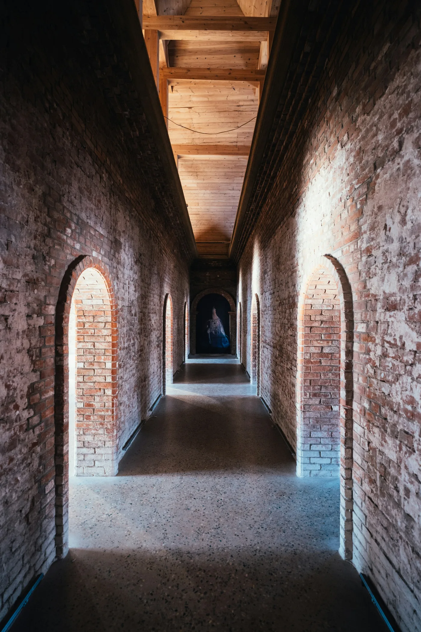 A long brick corridor with sunlit arched doorways and a timber ceiling.